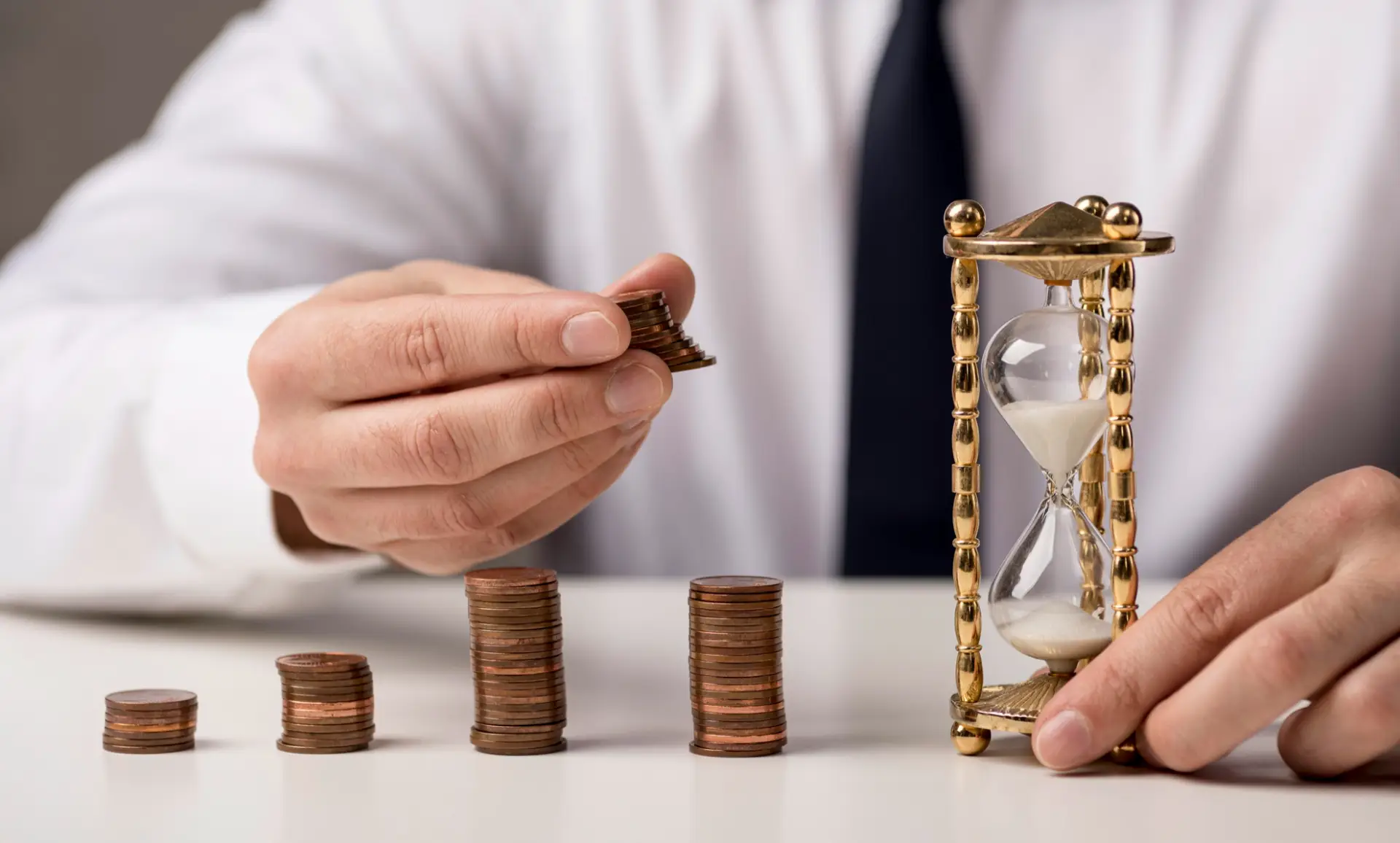A businessman stacks coins beside an hourglass, symbolizing the relationship between time and financial investment