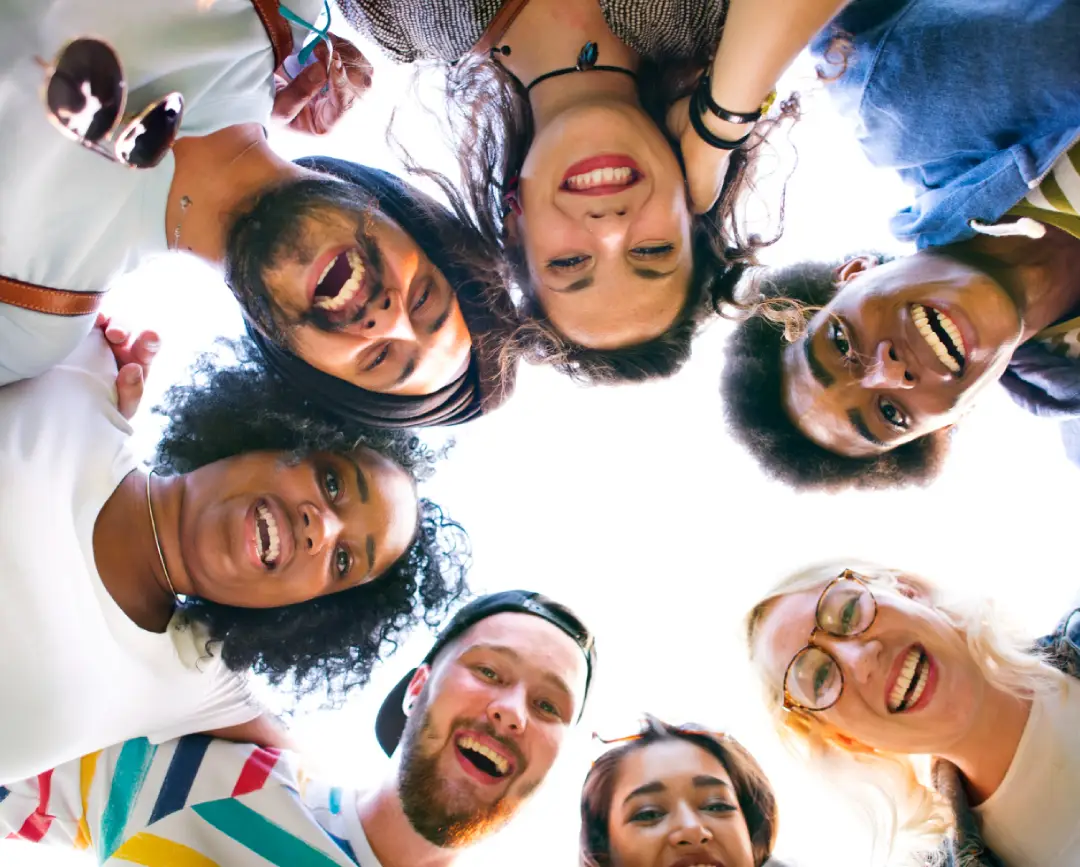 A group of diverse individuals gathers in a circle, looking down with smiles, illuminated by natural light from above