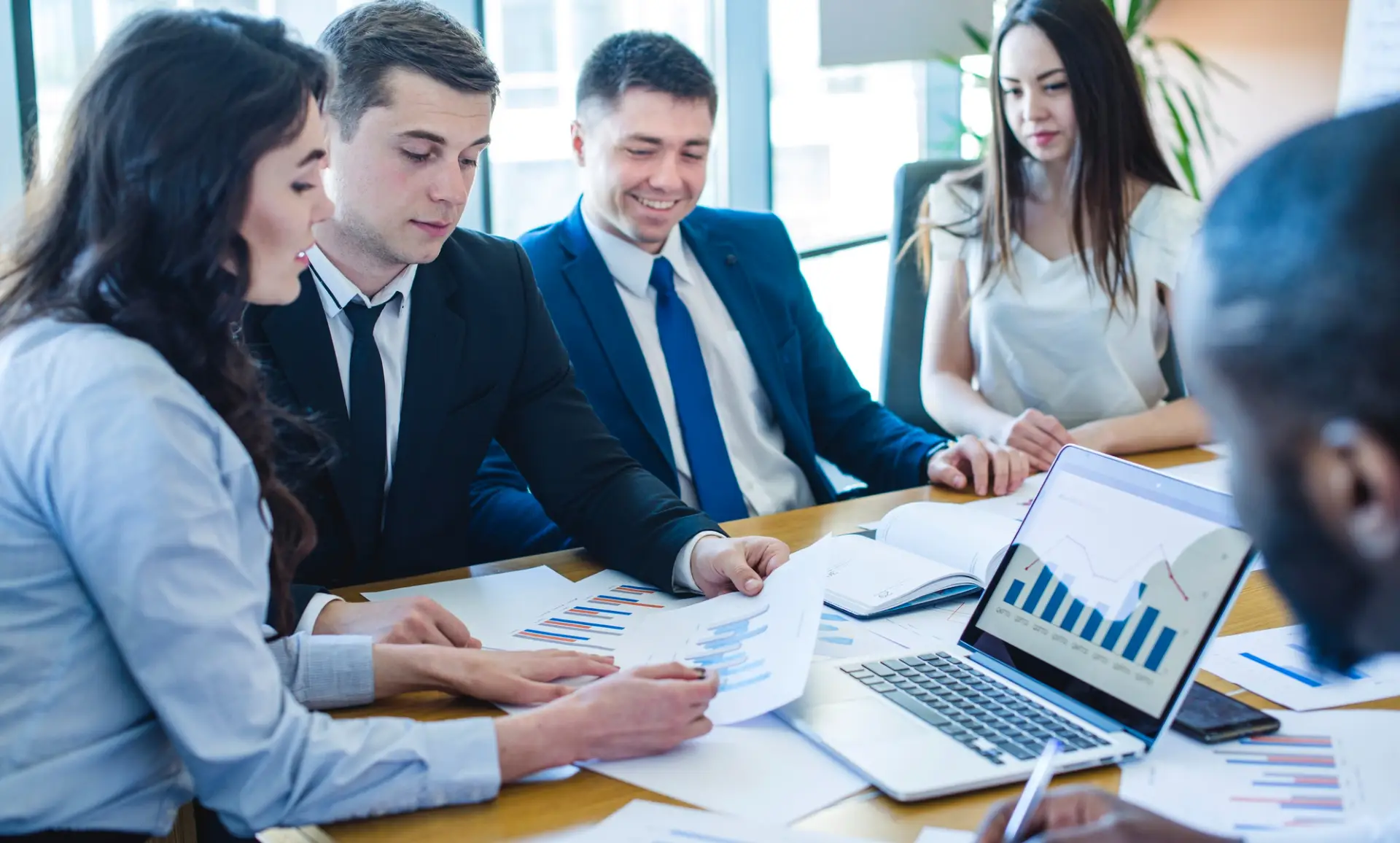 A group of professionals in business attire engage in a discussion around a table filled with charts and a laptop displaying data analyses